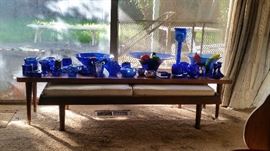 Beautiful cobalt blue candle holders and large bowls holding colorful glass fruits and vegetables. Also shown: mid-century low table and padded bench.