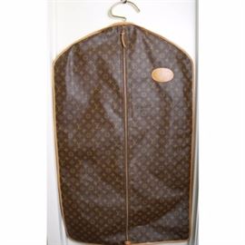 Louis Vuitton Garment Bag: A Louis Vuitton garment bag. This brown monogram canvas garment bag with tan leather trim has a hole in the top to accommodate a coat hanger, a central zipper, and a leather loop handle at the bottom. A round patch on the front of the bag is marked with the previous owner’s name and address, and a tag on the inside reads “Manufactured in the U.S.A. Under Special License to the French Co. Louis Vuitton; Vuitton and Fabric Design TM ® Vuitton et Fils, S.A. Paris.”
