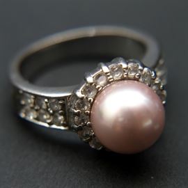Sterling Faux Pearl Ring With Rhinestones: A sterling silver ring with a central pink faux pearl. The faux pearl is surrounded by rhinestones that flow to its shank. It is marked “NVC” and “925.” The total approximate weight inclusive of nonmetal materials is 0.225 ozt.