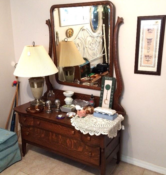 Antique oak lowboy dresser with tilt mirror. Brass accent table lamp with shade. Ottoman. Wall sconces with mosaic detail. Vintage lace doily. Vintage Avon soaps. Wall art. Scrolled cast metal headboard in white. Haeger pottery pedestal candle holder. Walking canes.