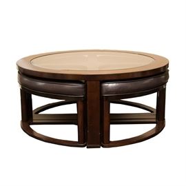 Glass Top Coffee Table with Stools: A glass top coffee table with stools. The table features a circular glass top set in a wooden frame with four legs and an X-stretcher. Includes four wedge-shaped stools with cushioned tops and wooden frames. Each piece has a brown finish.