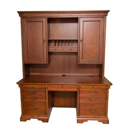 Wooden Office Desk with Built-In Power Controls: A wooden office desk with built-In power controls. The removable upper portion features a rectangular cornice and two cabinets flanking a center shelf with two drawers. A lamp between each cabinet top lights anything stored on the shelf. The bottom portion features a rectangular writing surface, two pedestals with three stacked drawers each, and a center pull-out keyboard tray. Faux drawer faces just below the top fold down to reveal built-in power control switches. Piece has a reddish-brown finish. All drawers feature dovetail joinery. All electronics worked upon testing.