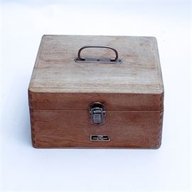 Wooden Japanese Medicine Box by Fujimoto: A wooden medical box from the company Fujimoto. This piece is made of wood and is in the tone of Japanese oak grain, features a simple handle and is secured with a metal clasp that has an equilateral cross on it; the universal symbol used in medicine. The interior is segmented into three slots, with a long top slot and two identical bottom slots. The maker’s mark is stamped on the front of the box just below the clasp.