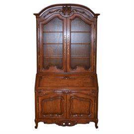 Auffray & Co. "Bressan" Louis XV Style French Country Secretary Desk: An Auffray & Co. “Bressan” Louis XV style French country secretary desk. This piece features a walnut finish. The top is arched and has a carved floral bouquet accent. A pair of doors with mesh wire fronts open to reveal four display shelves. Below the doors is a drop down door with a desk area within. There is a cubby area flanked by three drawers on each side, and a brown leather writing surface. Two cabinet doors below open to reveal additional storage space. The bottom edge is carved and scalloped, and the piece rises on cabriole legs ending in scroll feet.