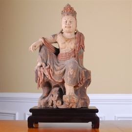 Wooden East Asian Statute: A wooden East Asian statute. This wooden figure, possibly a Bodhisattva, sits with one arm resting on the knee. The figure is painted with peach and gray paint, and rests on a wooden stand.