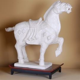 Plaster-Stone Horse Sculpture on Stand: A large horse sculpture on a stand. The white plaster-stone sculpture depicts a decoratively festooned horse with one leg raised and bent in the air. The sculpture is presented on a dark red and black stained wood Asian style stand.