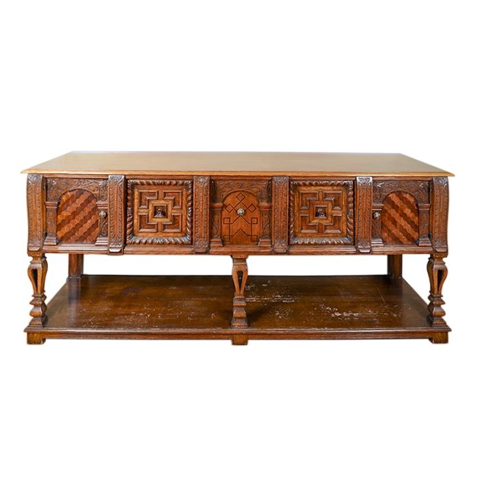 Eastlake Style Credenza: An Eastlake style credenza. This wooden credenza features a pecan finish with inlaid veneer along its front in a geometric array and carved sides. The front of the credenza includes two cabinets and two drawers, each featuring a silver tone or gold tone knob. The doors each feature intricate wood carvings and are separated by acanthus leaf applique. The lower half presents an open shelf with six baluster columns, which rest on six bracket feet.