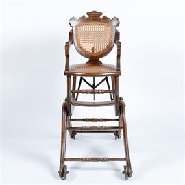 Antique Walnut and Cane Transforming Highchair in the Eastlake Style: An antique walnut and cane transforming highchair in the Eastlake style. This high chair features a carved rail over oval shaped cane back with carved stiles leading to carved arms, which are attached to an oval cane seat over a base which has turned ring stretcher support rails over a transforming base. The base has a conversion mechanism which with the folding out of a set of double bent legs, transforms the highchair into a child’s rocker; a set of wheels are attached for movement when converting.