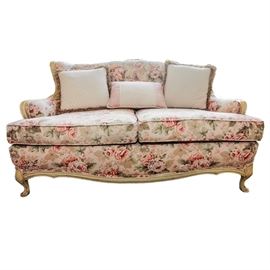 Vintage Floral Upholstered Sofa: A vintage sofa. Features an exposed blonde wood frame with carved floral detail to the top rail. It is upholstered with a colorful floral upholstery and includes three decorative pillows.