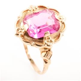 10K Yellow Gold and Faceted Pink Glass Cocktail Ring: A 10K yellow gold ring featuring one center faceted cushion cut pink glass stone mounted in flower shaped prong settings with a textured border, scalloped edges, and a carved diamond pattern to each side of the shank.