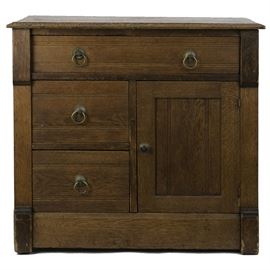 Antique Arts and Crafts Period Oak Commode: An antique arts and crafts period oak commode. This commode features one-over-two drawers along with a door to the right side, all with brass hardware and pegged construction. The sides exhibit column-like features with slightly overhanging corbels to top supporting the beveled surface. It is unmarked.