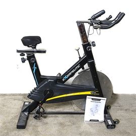 Exerpeutic Indoor Training Bike: An Exerpeutic indoor training bike. The upright stationary bike is black in color with the company logo in blue and yellow accents. The handles of the bike are cushioned in black. Comes with original manual.