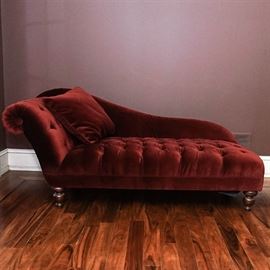 Tufted Red Chaise Lounge: A tufted red chaise lounge. This chaise lounge features a rolled side with slanting curved back, tufted red faux velvet upholstery and a matching square accent pillow. Chaise sits on turned wooden legs.