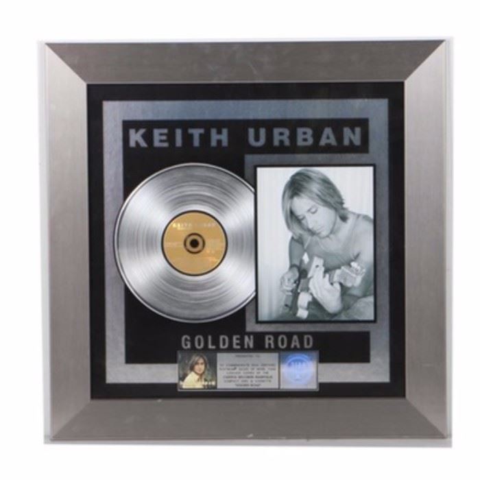 Keith Urban Platinum Record: A Keith Urban platinum sales award. Presented by the Recording Industry Association of America, this piece commemorates 1,000,000 copies sold of Urban’s album Golden Road with a metal nameplate at the base awarding it to Dann Huff. The piece is presented under glass in a silver tone metal frame with a wire wall hang to the back.