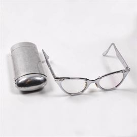 1950's Aluminum Cat Eye Glasses and Case: A pair of 1950’s aluminum cat eye glasses and case. Featuring a pair of lightweight metal framed eyeglasses with replaceable hinge lenses and offset nose rests. The stem of the glass frames is marked “5 3/4”.Also included is a coordinating metal case with hinged lid.