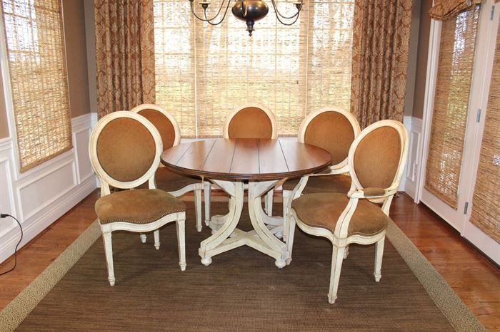 Round Pedestal Dining Table With Five Chairs: A French Country dining table with pedestal base and five French country dining chairs. The table features a distressed plank top finished in a medium stain. It is supported by a hand-painted pedestal base featuring curved legs, a low x-stretcher, and bun feet. This listing also includes two armchairs and three side chairs. Each features wooden frames constructed with balloon backs, curved seat rails, and square tapered legs. They are finished in a distressed hand-applied antique white and upholstered in a rich brown chenille with antiqued brass head trim. The armchairs feature padded manchettes.