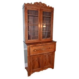 Antique Secretary Desk With Bookcase: An antique secretary desk with bookcase. This mahogany piece with rectangular top features a leaf and scroll carved pediment with beaded trim over two cabinet doors with glass panels and beaded trim work opening to three shelves. The bookcase sits atop a single drawer with scalloped trim and two knob handles that pulls out to additional drawers and storage space with green felt lined writing surface when drawer front is pulled down. The remainder of this piece features two paneled cabinet doors opening to two shelves over a scalloped apron terminating on bracket feet. There are no visible maker’s marks or labels on this piece.