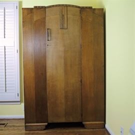 Art Deco Style Armoire Clothing Closet: An Art Deco style armoire clothes closet with lock. This tall cabinet is made of hardwood and oak veneer arranged in a contrasting grain. It features an arched pediment with ribbed molding and decorative panel at the center of the single door with period door pull and brass key. Inside is a brass closet rod with hooks on one end and three adjustable shelves added later in its life. Metal tag affixed to the inside of door reads “W Taylor upholsterer”. Located on the main floor.