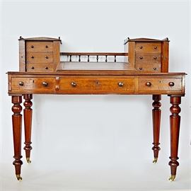 Antique Edwardian Dickens Style Mahogany Writing Desk: An antique Edwardian Dickens style cross-banded mahogany writing desk. This handsome desk desk is designed with three frieze drawers to each side of the top, the drawers constructed with hand cut dovetailed joints, with a center railed gallery between the drawers. The desk has a russet leather-covered slanted writing surface to the center, flanked by flat leather-covered writing surfaces to each side. The desk has round turned legs with polished end caps and casters.