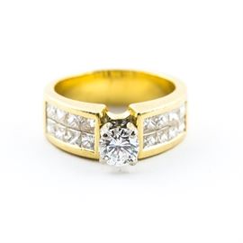18K Yellow Gold and Diamond Ring: An 18k yellow gold and diamond ring. It features a round brilliant cut center stone set on four prongs, and two columns of princess cut side diamonds on the golden band.