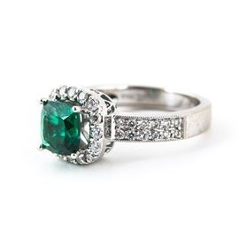 18K White Gold, Emerald, and Diamond Ring: An 18k white gold, emerald, and diamond ring. It features a synthetic emerald center stone surrounded by a hoop of diamonds around the perimeter. Two rows of diamonds cascade down the shoulders of a white golden band.