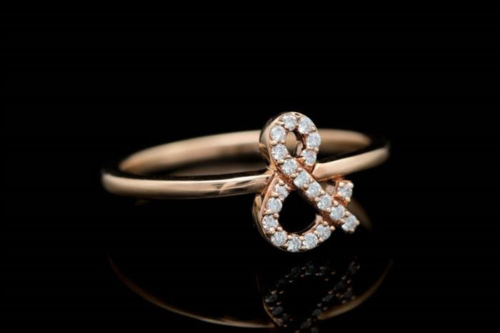 18K Rose Gold and Diamond Ampersand Ring: An 18K rose gold and diamond ampersand ring. The rose gold ring features an ampersand lined with twenty diamonds.
