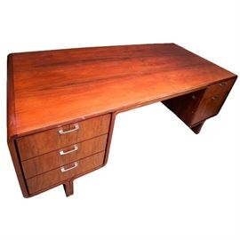 Mid Century Modern Rosewood Executive Desk: A Mid Century Modern executive desk. This desk features rosewood veneers throughout. The front of the desk features a kneehole flanked by three drawers on one side and two graduated drawers on the other; all have silver tone pulls. The opposite side of the desk features a large rectangular panel with rounded corners, all in rosewood veneers. It sits on two legs extended from the frame of the desk.