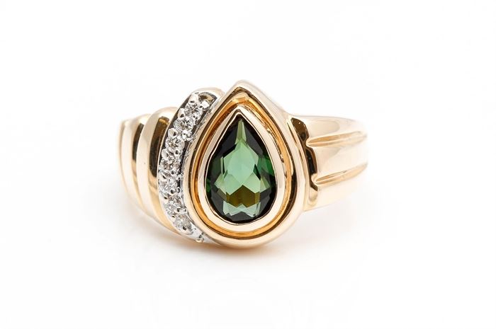 14K Yellow Gold Green Tourmaline and Diamond Ring: A wide asymmetrical yellow gold shank holds a bezel set faceted pear cut green tourmaline to the center and a half halo of round cut diamonds to one side.