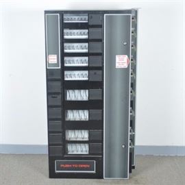 Vending Machine: A vending machine. This vending machine is black and silver toned. Along the right side are slots for coins that turn to select the item. The front of the machine has selection windows. Included is a coin box. Marked “Antares Corporation" and serial no: A-27425.