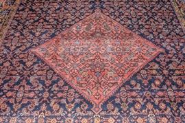 Red and Dark Blue Herati Pattern Handwoven Area Rug: A red and dark blue Herati pattern hand woven area rug. The large rug features a red diamond shaped medallion with arrows at each corner, inside a navy blue pentagonal field with a toothed edge, and red spandrels. Hereti designs in shades of light blue, red, and yellow fill the space inside the medallion and field. The dark blue main border and yellow guard borders depict carnation designs The rug is finished with a dark blue side cord on the longer side and white fringe at the shorter ends. No tag.