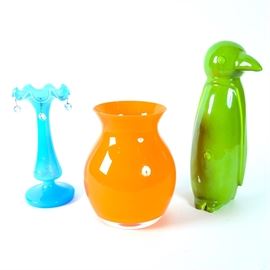 Colorful Glass Vases and Ceramic Penguin Figurine: A group of colorful vases and a figurine. Included is an antique blue glass vase with a ruffled rim and hanging octagonal prisms. Unmarked, the vase at one point had gold tone designs painted on its exterior. Also included is an orange glass vase and a green ceramic penguin figurine, both of which are unmarked.