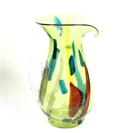"Blenko" Hand-Blown Art Glass Pitcher: A “Blenko” hand blown art glass pitcher. Featuring a twelve-inch tall pitcher with a pour spout in green and multi-colored glass. “Blenko 004” is etched into the bottom side of the pitcher and there is a sticker attached that reads “Blenko Handcraft Made in USA”.