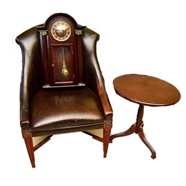 Bombay Company Chair, Accent Table, and Clock: A Bombay Company chair, accent table, and clock. This three piece set includes a walnut stained circular accent table with a tripod base, a wooden rectangular pendulum hanging wall clock with gold tone metal hands and trim and a brown faux leather chair sitting on tapered carved wood legs. Each piece is stamped with “Bombay Company” to the underside.
