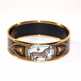 Hermès Grand Apparat Bracelet: A Grand Apparat bracelet by Hermès of Paris. The bracelet has gold tone metal construction with an enameled equestrian design. It features a burgundy enamel ground with a pattern of horses and swags in white and gold tone. The bracelet is marked “Hermès Paris,” and “made in Austria” on the interior. It measures 2.75" in diameter and 8" in circumference. It is 1" wide.