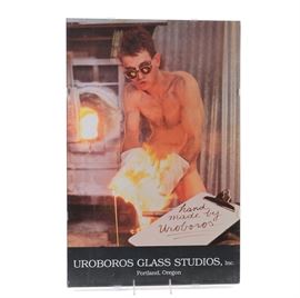Uroboros Glass Studios Offset Lithograph Poster: A Uroboros Glass Studios offset lithograph poster. This piece exhibits a photograph depicting a naked man wearing sunglasses while working with flaming hot glass. Superimposed to the original photograph is the image of a clipboard displaying the words “hand made by Uroboros.” The lower margin is labeled “Uroboros Glass Studios, Inc. Portland, Oregon.” This piece is displayed behind glass, held together with plastic brackets; with strings located at the top and bottom center for hanging.