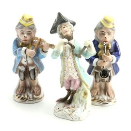 Porcelain Colonial Attire Monkey Musician Figurines: A selection of three porcelain monkey figurines wearing Colonial period attire while playing musical instruments. They are hand-painted in bright colors with gilt accents, and include a violinist and a saxophonist, wearing yellow hats. The third monkey in the green tailcoat with a black tricorn hat is crafted to resemble and is styled after the Meissen Monkey Band and bears an unknown maker’s mark to the underside.