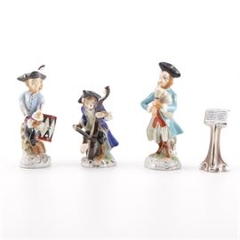 Colonial Attire Monkey Musician Figurines With Podium: A selection of three porcelain monkey figurines wearing Colonial period attire and tricorn hats while playing musical instruments. There is also one podium. They are hand-painted in bright colors with gilt accents and include a bagpipe player, a cellist, and a drummer. They are crafted to resemble and are styled after the Meissen Monkey Band and bear an unknown maker’s mark to the undersides.