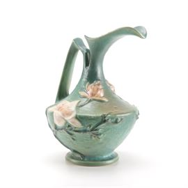 Roseville "Magnolia Blue" Ewer: A Roseville pottery Magnolia Blue ten inch ewer. The ewer has a defined lip and a dip before the angular handle which extends out, then down to connect to the body. This ewer is blue with warm accents and magnolia flowers along the stem and body one as a bud and the other in full bloom with branches around the base. The bottom is marked “Roseville.”