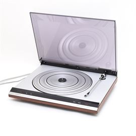Bang & Olufsen Beogram 2404 Turntable: A Bang & Olufsen Beogram 2404 record player. Includes a DIN to RCA gold plated adapter. Features a smoked plastic top with a silver tone deck, arm and turntable. The case is finished in faux wood, silver tone metal and black plastic. The player powered on when tested, no further testing was done.