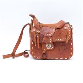 Handmade Tooled Leather Western Saddle Handbag: A handmade tooled leather western saddle handbag. Features a tooled leather saddle style with embroidered suede seat. The leather is finished in a natural london tan with cream tone whipstitching and silver tone hardware throughout. The bag top is lined in sheepskin. It is not hallmarked.