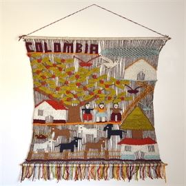 Woven Folk Art from Colombia: A folk art style woven textile wall hanging from Colombia. This handmade piece is decorated in green, red, blue and orange threads with hints of white and black. It sits atop a medium brown tone backdrop with “Colombia” woven in brown at the top. It hangs by a twisted top thread and is perfectly finished at the bottom with multi-color thread fringe.