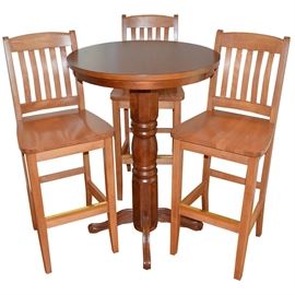 Pub Table and Chairs: A round pub table and set of three bar chairs. The table sits atop a turned wood pedestal base with four curved, fluted feet. Chairs are mission style and presented in a maple finish with straight legs and stretchers. Tag below seating reads “Made in Canada by Holsag Canada Lindsay, Ontario.” A matching pub table and chair set is available, item 17CIN187-011.