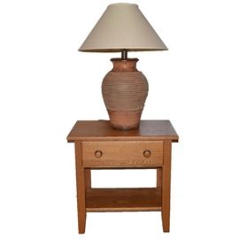 Mission Style Oak End Table and Pottery Table Lamp: A mission style oak end table and a pottery table lamp. The oak table includes a shallow storage drawer with matching wood and antique brass tone round knobs. The sides feature mission style wood stretchers that attach to an open lower storage shelf. The table lamp is made of pottery and shaped as a vase with beautiful texture highlighting the body. Lamp includes an ivory tone fabric bell shade.