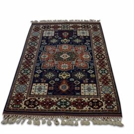 Hand-Knotted Kazak Area Rug: A hand-knotted Kazak area rug. This rug features a central medallion in green, light blue, yellow, and cream over a deep blue backdrop. Smaller square medallions appear at each of the center’s corners in cream, red, and light blue. A pattern of colorful, geometric floral motifs make a border on a cream ground color. Cream colored, knotted fringe appears on either end. There is a label attached to the rug.