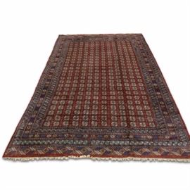 Hand-Knotted Wool Bokhara Palace Rug: A hand-knotted wool Bokhara palace rug. This rug features a repeating pattern of “elephant foot” guls on a red backdrop. The resolved border is made up of additional guls in single file on a red ground color. Cream colored, wool fringe appears on either end of the rug. A label is attached to the underside with details about the rug.