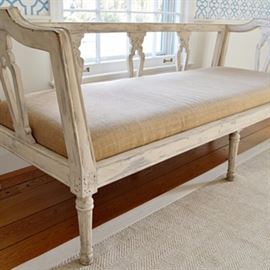 French Provincial Bench: A French Provincial bench. This sofa style bench has a white washed wood frame and natural jute upholstery. There is engraved floral detailing along the supports. It sits on turned legs and includes a white padded cushion.