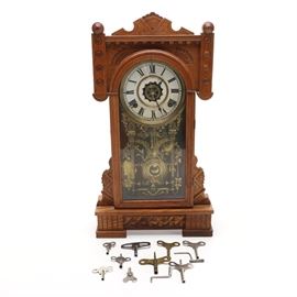 Antique Eastlake-Style Mantel Clock With Keys: An antique mantel clock with keys. This Eastlake-style clock is housed in a golden oak frame. The dial reads with Roman numerals. Clockworks are visible behind a glass panel that has been decorated with gold tone Roman columns and scrolling vines and acanthus. The lot includes eleven metal clock keys of various styles and sizes. The clock functions and chimes but has not been tested for accuracy.