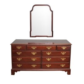 Davis Cabinet Company Dresser With Wall Mirror: A Davis Cabinet Company dresser with a wall mirror. This solid wood dresser has a rectangular top with a beveled edge. It features nine drawers with brass tone, drop-handle pulls. The dresser has a shaped skirt and rests on four feet. Both the dresser and the frame of the wall mirror have a cherry lacquered finish. A placard to the interior of one of the drawers reads “Davis Cabinet Company, Craftsmen of Fine Solid Wood Furniture, Nashville, Tennessee”.