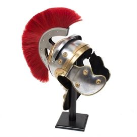 Reproduction Roman Centurion Helmet: A reproduction Roman Centurion helmet in stainless steel and brass. This distinctive helmet features a bright red plume and sits upon a black wooden stand.
