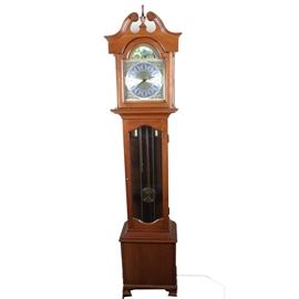 Daneker "Cumberland" Grandmother Clock: A Cumberland grandmother clock by Daneker. This clock is made of a dark mahogany with gold tone accents and features a broken arch pediment and finial. The clock has an ornate gold tone face with black Roman numerals and filigree accents under a matching moon dial. The front glass displays a gold-tone pendulum.